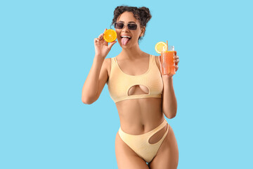 Young African-American woman in swimsuit with cocktail and citrus showing tongue on blue background