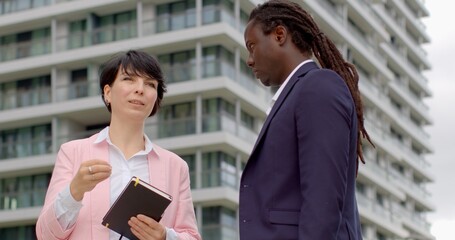 Woman real estate agent talking to male client against backdrop of modern buildings. Concept of property consultation, urban real estate, and professional communication.