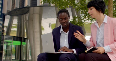 Using laptop with artificial intelligence, businesspeople engages in remote work, efficiently handling tasks and responsibilities from location with internet access. Afro man and white woman talking.