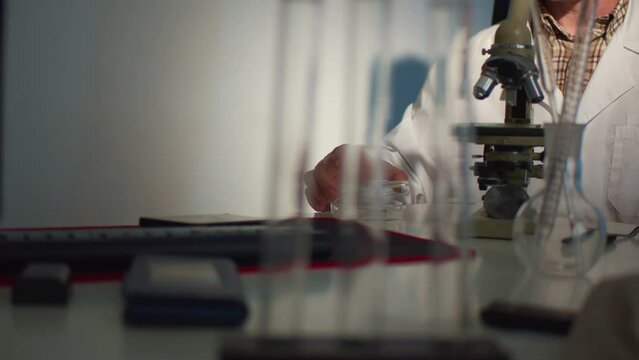A male student researcher in glasses and a white coat at his desk in a work environment uses a microscope to study an insect