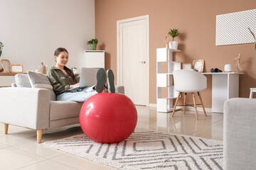 Young Asian woman with fitball using laptop on sofa at home