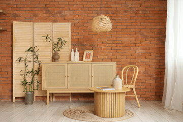 Wooden chest of drawers, table, chair and folding screen near brick wall in stylish interior of...