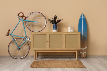 Surfboard, bicycle and wooden chest of drawers with flowers near beige wall