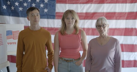 Three voters, two women and one man, smile while standing in front of giant United States flag, on wall in background