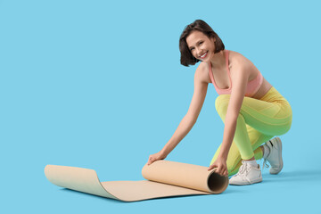 Sporty young woman rolling out fitness mat on blue background