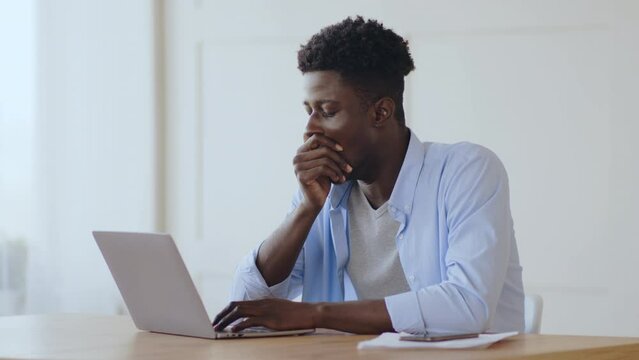 Overwork concept. Tired african american guy yawning at workplace, typing on laptop at home