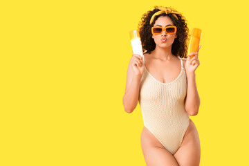 Beautiful young African-American woman in swimsuit with bottles of sunscreen cream on yellow...