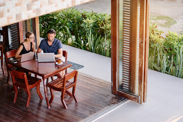 Coworking and freelance concept. Young  bearded man  and young woman working together on laptop computers while sitting on cafe terrace.