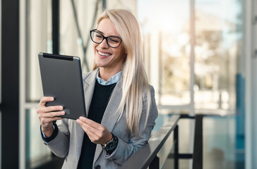 Fototapeta premium Smiling blonde female IT specialist working on digital tablet while standing in a lobby of an office building. She is wearing business clothes and eyeglasses.