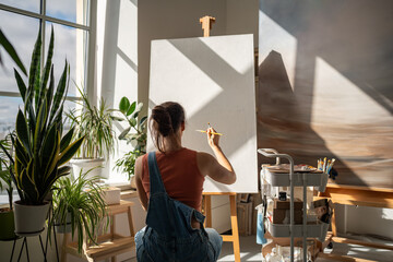 Creative artist woman starts to draw artwork on new white canvas on easel with brush at equipped...