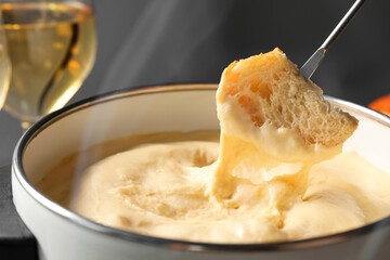 Dipping piece of bread into fondue pot with melted cheese on grey background, closeup