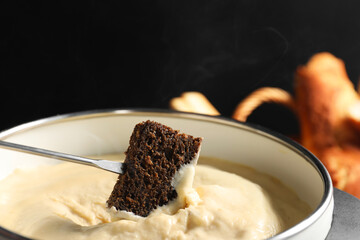 Dipping piece of bread into fondue pot with melted cheese on black background, closeup