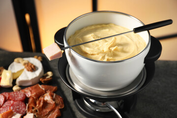 Fork with piece of ham, fondue pot with melted cheese and snacks on table, closeup