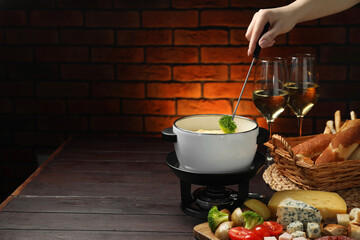 Woman dipping piece of broccoli into fondue pot with melted cheese at wooden table with wine and...