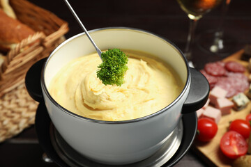 Dipping piece of broccoli into fondue pot with melted cheese at table with snacks, closeup