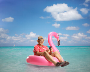 Mature male tourist sitting on a famingo swimming ring in the sea and using a mobile phone