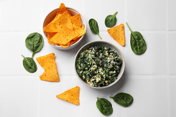 Tasty spinach dip with egg in bowl and nachos chips on white tiled table, flat lay