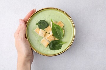 Woman with bowl of delicious spinach cream soup on light grey table, top view