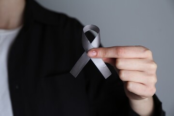 Woman holding grey awareness ribbon on color background, closeup