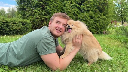 Young man is lying on green grass in park with his cute little Pomeranian Spits dog, guy play, hug...