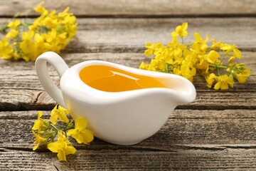 Rapeseed oil in gravy boat and beautiful yellow flowers on wooden table, closeup