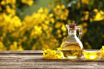 Rapeseed oil in glass jug, bowl and beautiful yellow flowers on wooden table outdoors, space for...