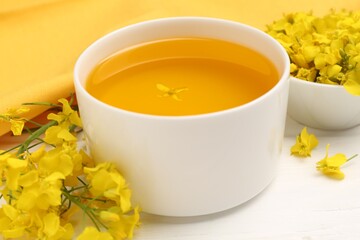 Rapeseed oil in bowl and beautiful yellow flowers on white wooden table, closeup