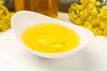 Rapeseed oil in gravy boat and beautiful yellow flowers on white wooden table, closeup