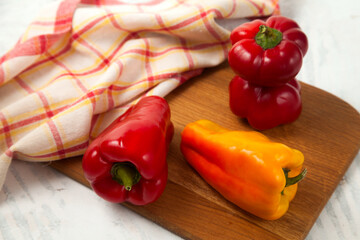 Cutting board with several whole red and yellow bell pepper and red kitchen towel on white wooden background. .