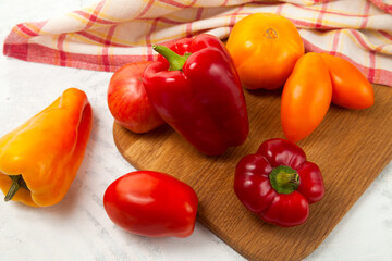 Cutting board with several red and yellow tomatoes and bell pepper on white wooden background with red kitchen towel..