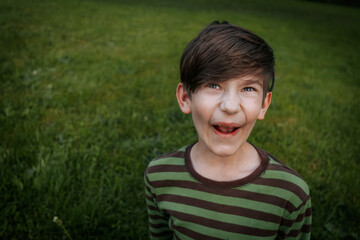Obraz premium young boy with brown hair and blue eyes wearing a green and brown striped shirt makes a funny face while standing on a grassy field. The background is blurred