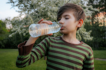 young boy drinking water from a plastic bottle, wearing a striped shirt, with a blurred green outdoor background.