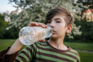 young boy drinking water from a plastic bottle, wearing a striped shirt, with a blurred green outdoor background.