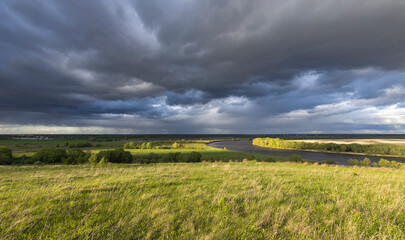 A serene meandering river flows through a vast grassy field under a dramatic sky filled with dark storm clouds and patches of light.