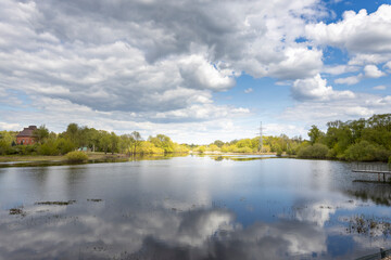A serene lakeside scene with a calm water surface reflecting the sky and greenery, featuring a wooden pier and walkway, surrounded by trees and electricity poles