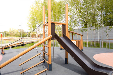 An outdoor playground featuring a wooden climbing frame with a slide, rope ladder, and swing set,...