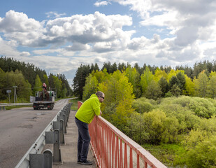 A solitary man in a yellow jacket and sunglasses stands on a bridge, gazing over the railing at a...