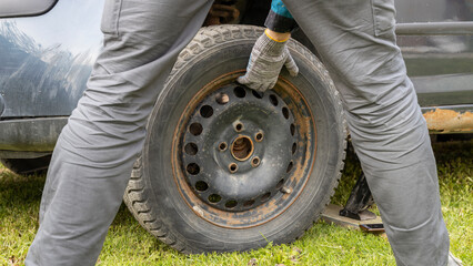 A man wearing gloves changes a car tire on the grass, holding a wheel removed from the car's hub...