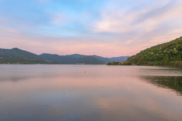  A serene sunset over a tranquil lake with rippled water, mountains in the distance, and a clear sky with soft clouds