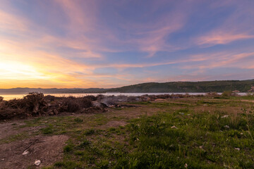  scene at the edge of a water body during sunset or sunrise, with a pile of burning logs and scattered garbage, highlighting the juxtaposition of natural beauty and environmental decay...