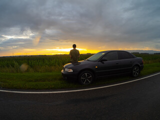 A young man sits on the hood of a car and looks at the sunset over a field in the rain. Traveling by car