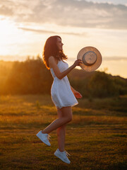 A happy young woman in a dress and a straw hat poses and jumps in a sunny field.