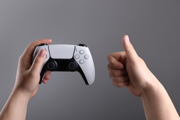 Woman using wireless game controller and showing thumbs up on grey background, closeup