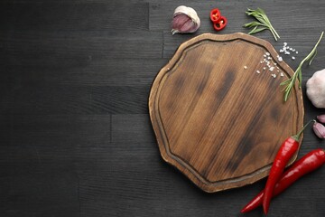 Cutting board, garlic, rosemary and chili peppers on black wooden table, flat lay. Space for text