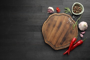 Cutting board, spices, garlic, rosemary and chili peppers on black wooden table, flat lay. Space...