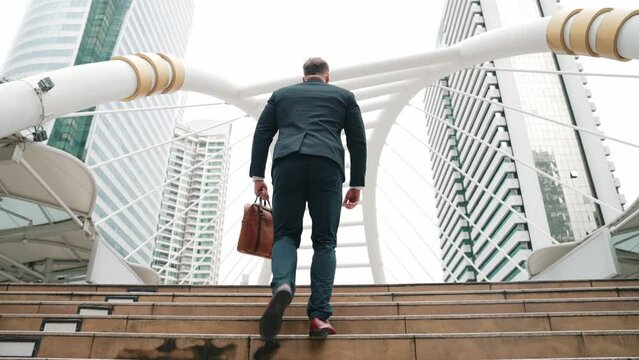 Smart caucasian businessman walking up stairs surrounded by urban view. Back view of ambitious project manager finding challenging job or investment opportunity at city with modern building. Urbane.