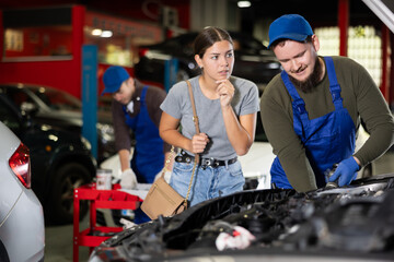 Young guy mechanic advises young woman client on repairing under hood of car in car service station