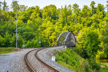 A curved railway bridge in Rataje surrounded by dense greenery and trees during summer, with railway tracks leading up to the bridge. - Powered by Adobe