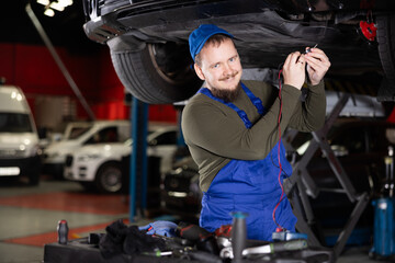 Skilled male auto mechanic in blue overalls measuring capacity of automobile detail in car workshop