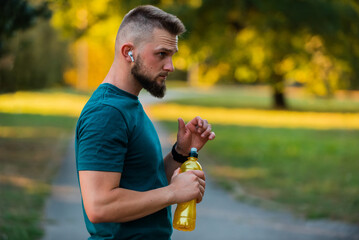 A young fit man listening to music on wireless headphones and drinking water or nutrition juice...